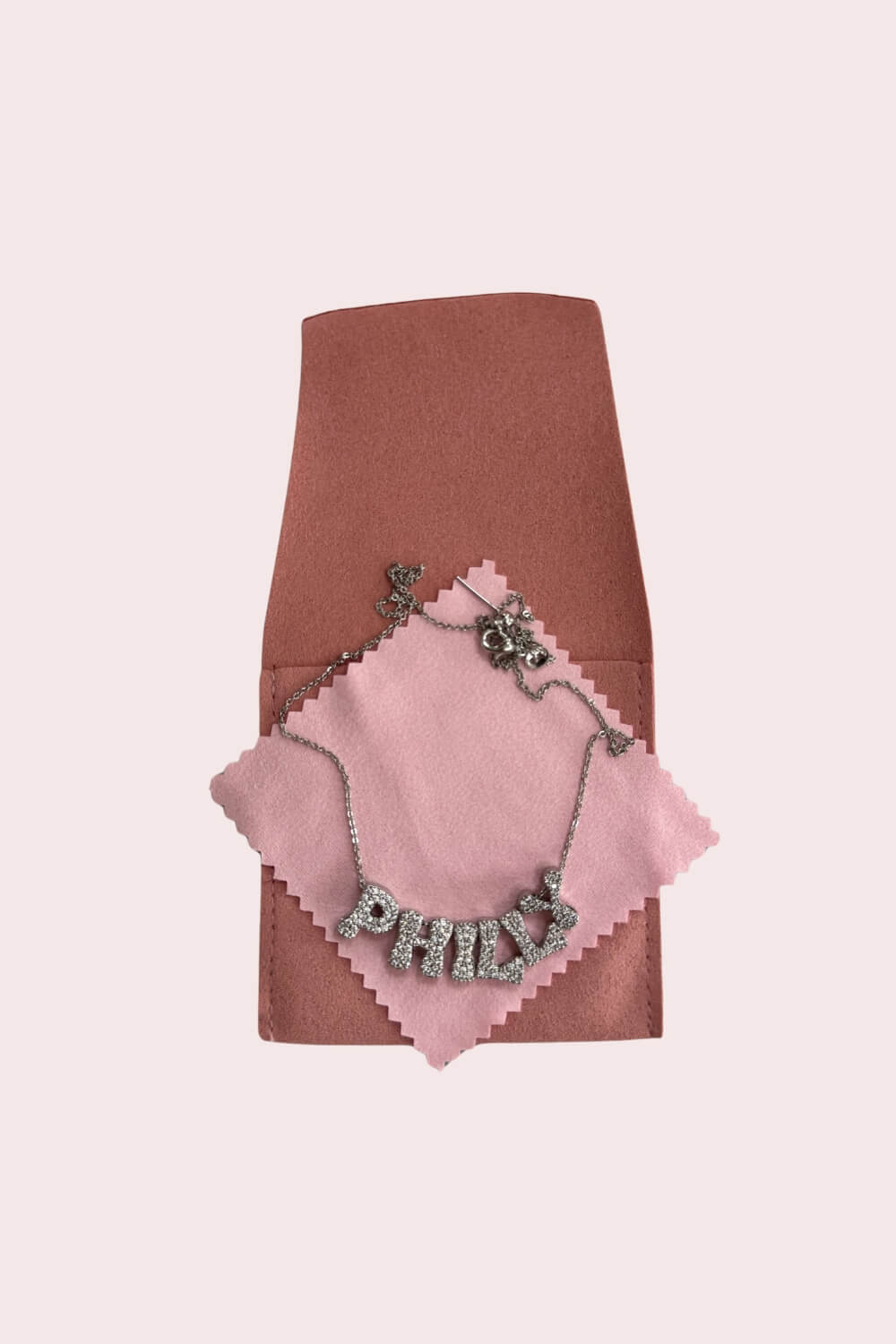 Necklace with 'PHILLY' in pave letters, on a pink cloth and brown protective bag.